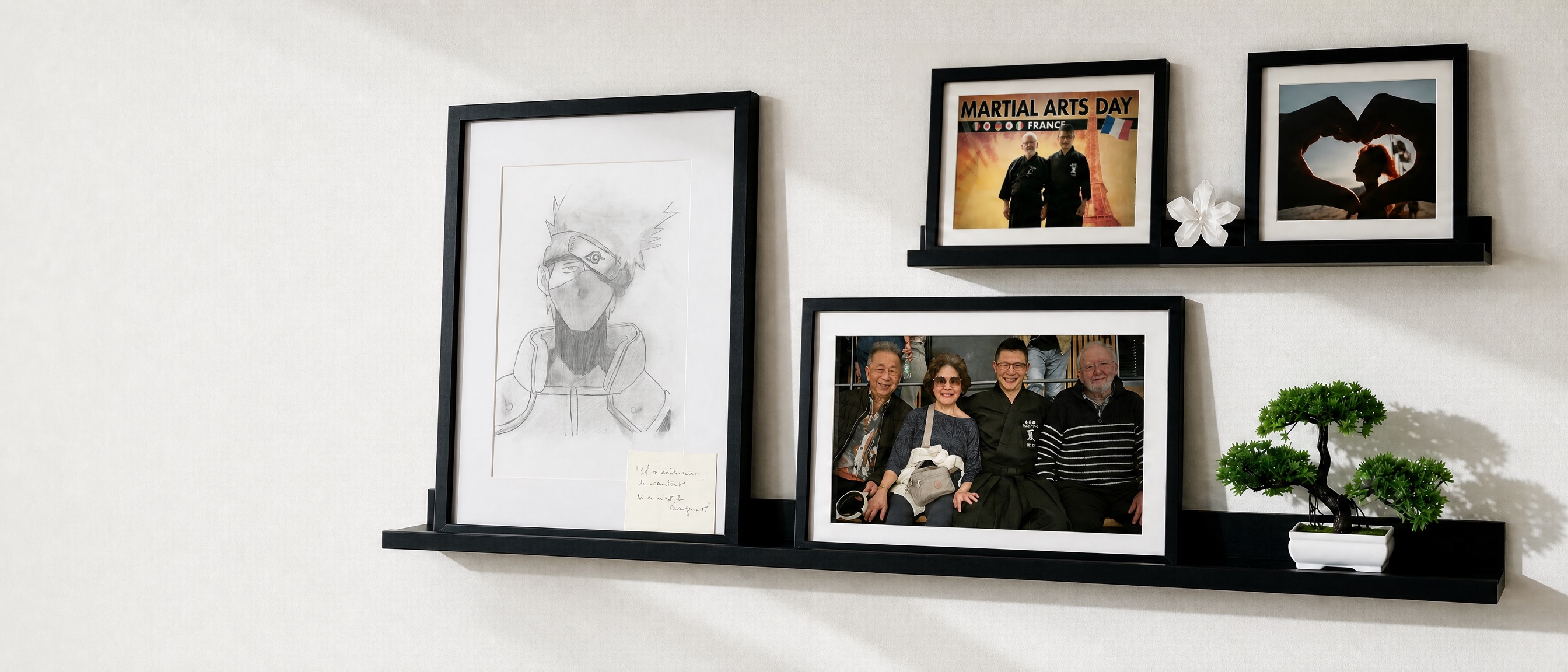 Un mur blanc conduit vers deux étagères Mosslanda présentant le dessin de Kakashi réalisé par Khey-leng, les photographies de la famille de Tex, de Guy Sensei et de Lyse, accompagnées d'un origami blanc et d'un bonsaï.